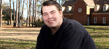 young man smiling and sitting outdoors with trees and a brown house